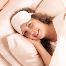 Woman lying laughing on rosé satin pillow – white Peace Silk sleep mask pushed up on forehead